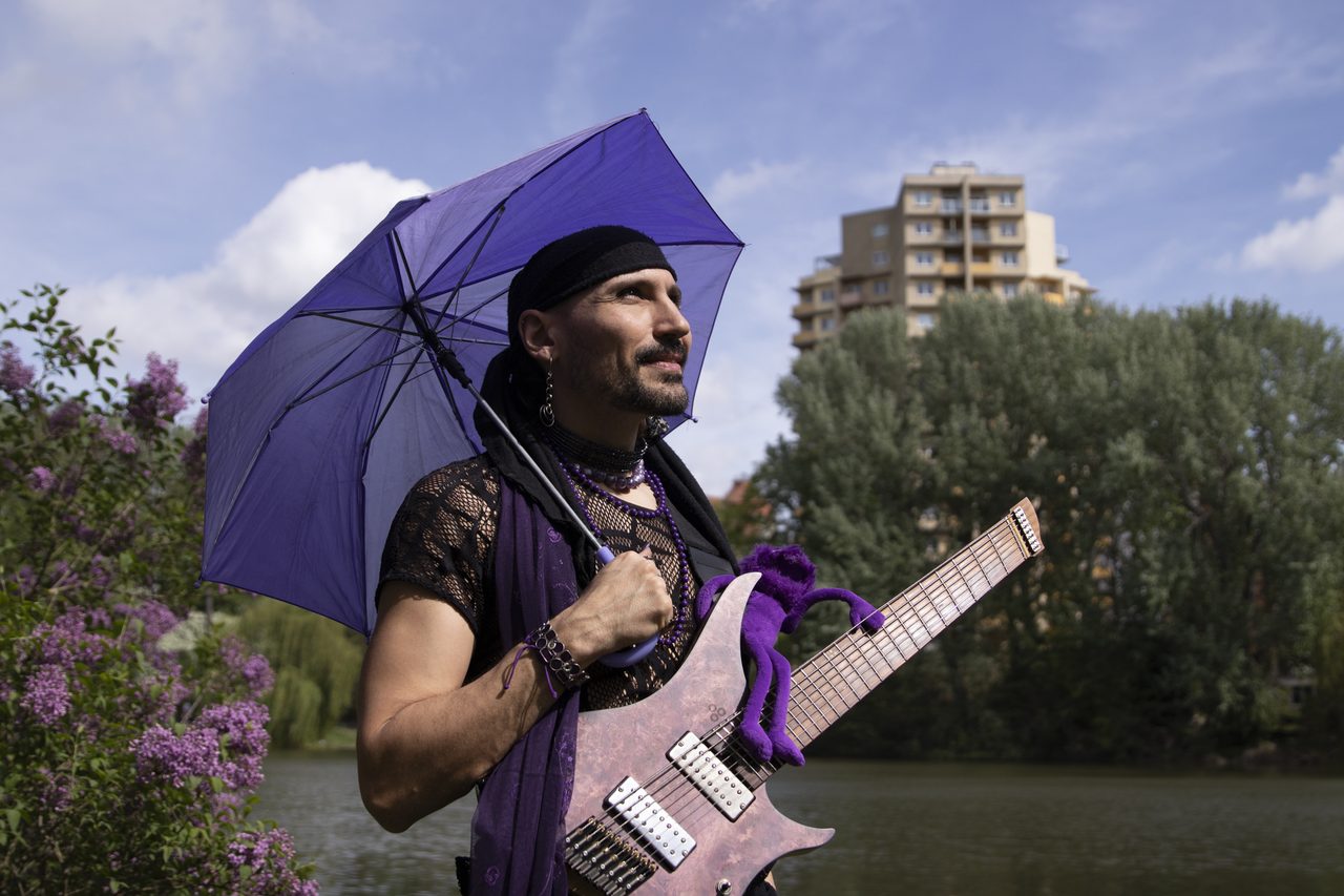 Danijel Milosevic holding a purple umbrella and a guitar with a purple plush toy on it, standing by a lake with blooming lilacs